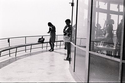 A balcony view features two women in what appears to be a seaside resort. One woman, in a dress, speaks into a phone. Another...