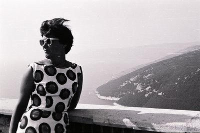 Elegant woman in a white, spotted dress and dark sunglasses poses overlooking a coastal vista. The dramatic, sweeping landsca...