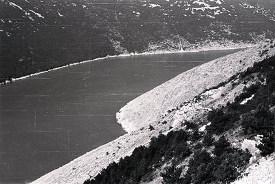 High-angle view of a coastal landscape featuring a calm body of water meeting a steeply inclined, rocky shoreline covered in ...