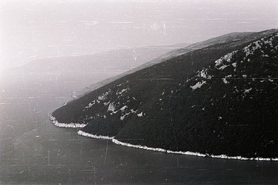 High-angle, monochrome aerial view depicts a coastline, likely the Black Sea. Rugged, forested slopes descend to a rocky shor...