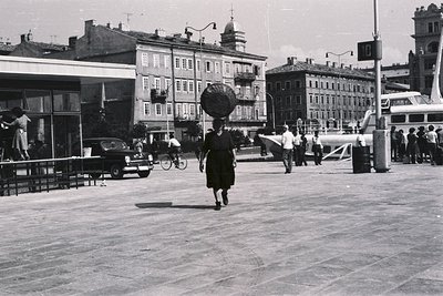 A woman carries a basket on her head, traversing a paved plaza. Buildings with tiered facades rise in the background, featuri...