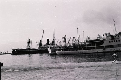 Three cargo ships docked in a harbor, viewed from a stone promenade. Multiple cranes are visible along the ships’ decks. A fi...