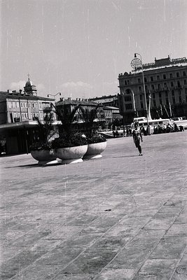 A view of a paved square with prominent, rounded planters holding foliage. Classic architecture & a large hotel ("Hotel Splen...