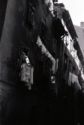 A black and white photograph showcasing a densely packed urban scene. Laundry hangs from lines strung between buildings, reve...