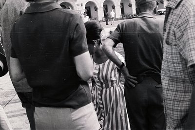 A view through the backs of several men, one wearing a military-style uniform, focuses on a woman in a vertically-striped dre...