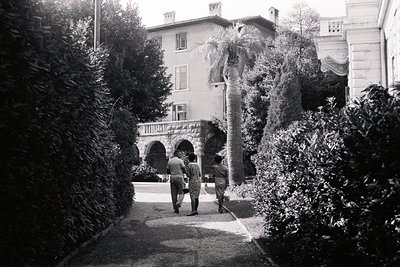 Classic mid-century architectural scene: two figures stroll along a path toward a grand villa with arched porticos & ornament...