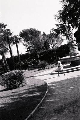 Elegant woman in a dress stands near a classical fountain within a lush, manicured garden. A stately statue adorns the founta...