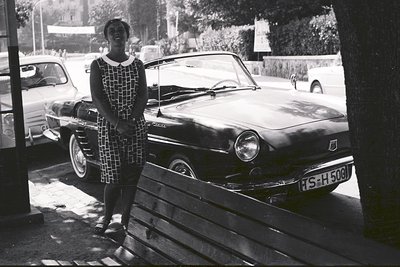 A young woman stands next to a dark-colored, open-top car, likely a Renault Floride, bearing a license plate "TS-H-508". She ...
