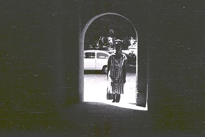 A woman in patterned dress stands in a courtyard, framed by an arched doorway. A vintage van is visible beyond. Likely 1960s ...