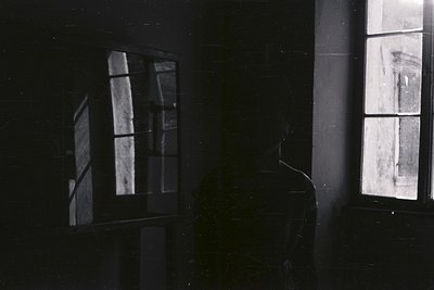 A solitary figure stands with their back to the camera, positioned near a window with gridded panes. Strong sunlight illumina...