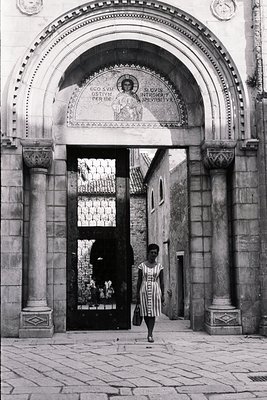 Monochromatic image depicts a woman walking through an ornate stone archway. The arch features a central mosaic of a religiou...