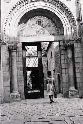 A monochrome photo captures a woman departing through an ornate archway, potentially a church entrance. Inscribed Latin text ...