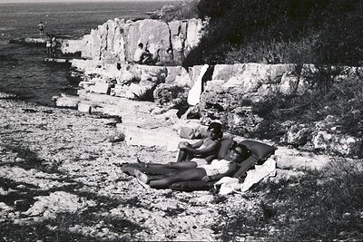 Two individuals lounge on a patterned blanket amidst rocky terrain near the seaside. A stone cliff face rises sharply behind ...