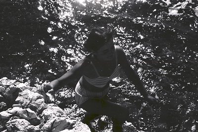 A woman, clad in a patterned swimsuit, ascends a rocky incline; sun-dappled water creates a shimmering effect overhead. The b...