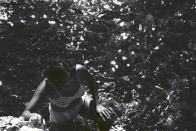 A monochromatic view captures a person partially submerged in shallow water, likely at a natural spring or rocky pool. Detail...