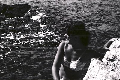 A woman in a bikini looks out over a rocky, seaside landscape. Black and white photography suggests a mid-20th century time p...