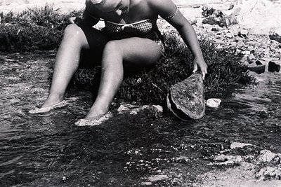 A woman in a patterned bikini sits on rocks in a shallow stream, examining a large, flat stone. The scene appears naturalisti...