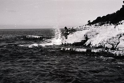 A person leaps from a rocky cliff into the sea, creating a splash. Textured limestone formations define the shoreline. Severa...