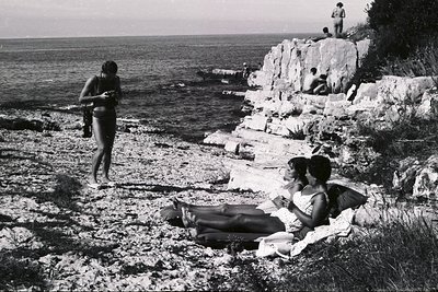 Coastal scene: Several figures relax on rocky bluffs overlooking the sea. Two young women lounge on pillows; a third figure s...