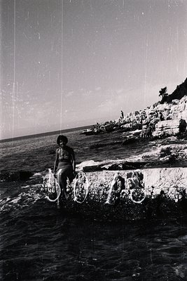A person sits on a stone quay, partially submerged in water, overlooking a seascape. The rocky coastline rises on the right. ...