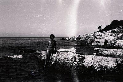 A woman in a vintage swimsuit sits poised on tiered rock formations along a rocky coastline, waves crashing nearby. Likely ta...