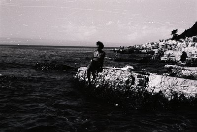 A young man sits perched on a rocky outcrop overlooking the sea. Textured stone steps ascend the cliff face in the background...