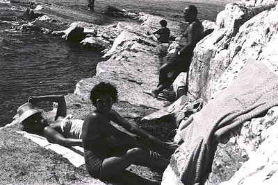 B&W scene captures a rocky coastal landscape with three people enjoying a day at the seaside. Two women in swimwear and a man...