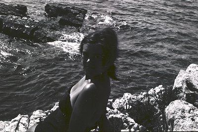 A young woman sits on a rocky coastline, gazing towards the sea. Sharp contrasts suggest bright sunlight & a vintage photogra...