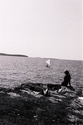 A lone child sits on a rocky outcrop overlooking a vast body of water, likely a lake or sea. A person flies a kite in the dis...