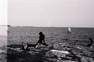 B&W image captures a seaside scene with figures silhouetted against the water and sky. Two figures are visible near the shore...