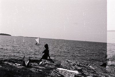 A lone figure sits on rocks overlooking a wide, still body of water, likely a bay or inlet. A single sailboat is visible on t...
