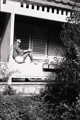 A stylish man, sunglasses on, relaxes on a low wall within an outdoor space. He wears a striped shirt and white shorts. Archi...