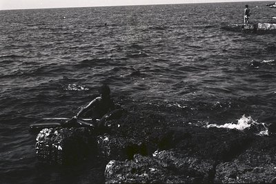 Monochrome image depicts a coastal scene. A person sits on a rocky outcrop, partially submerged in water, with another figure...
