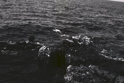 A lone figure swims in turbulent, dark water near rocky shoreline. The monochrome image emphasizes the texture of the waves a...