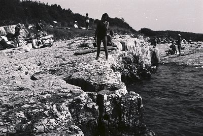 A young woman in a dark swimsuit stands on a rocky ledge overlooking the sea. Several figures are scattered across the beach ...