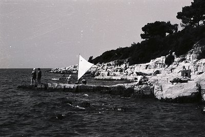 A group utilizes a makeshift sail attached to a frame on a stone quay jutting into the sea. Rocky coastline rises steeply, co...