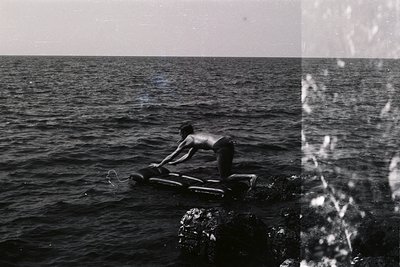 A young man kneels on an inflatable raft amidst a dark, expansive seascape. Rocks jut from the water's surface. The photograp...