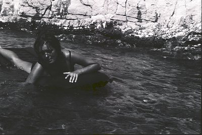 A young woman lies partially submerged in shallow, clear water, with a rocky cliff face visible in the background. She appear...