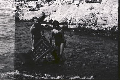 Two figures, a man and a woman, wade through shallow water at a rocky coastline, carrying an inflatable raft. Visible landsca...