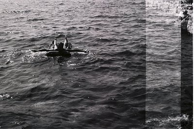 A swimmer floats supine in turbulent, dark water, arms outstretched. A blurred figure sits on rocks to the right, overlooking...