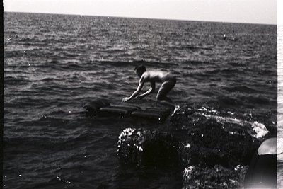 A young man, appearing nude, leaps from a rocky outcrop into the sea. The image captures a moment of action with blurred moti...