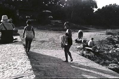 A candid black-and-white street scene. Two women in vintage swimwear stroll along a paved walkway. Others relax in the backgr...
