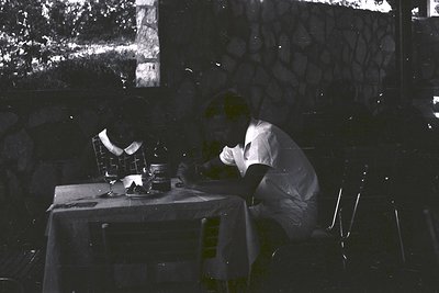 Man seated at an outdoor table covered with a white tablecloth, seemingly relaxed and contemplative. Stone architecture and g...