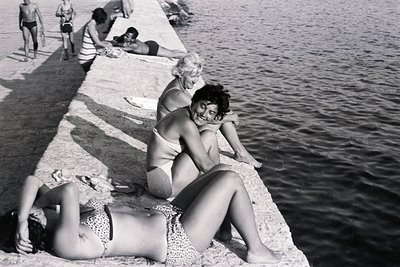 B&W image depicts sunbathers relaxing on a concrete retaining wall alongside a dark water body. Two women, one reclining & on...