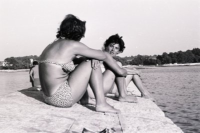 Two women in vintage swimwear sit on a stone embankment overlooking a seaside, likely in Bulgaria. The woman on the left wear...