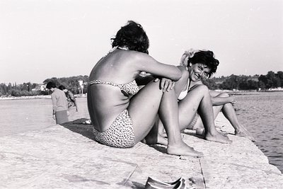 Two women in swimwear seated on a concrete retaining wall overlooking a beach, likely enjoying a seaside resort. A third figu...