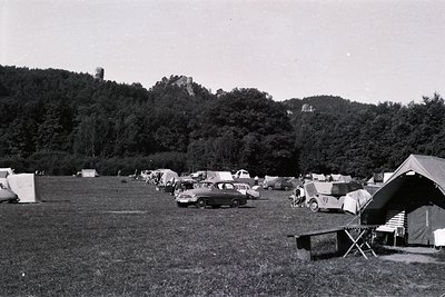 A grassy field hosts a vintage car rally/camping event. Several classic cars and numerous tents dot the landscape, backed by ...