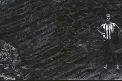 Striking monochrome image. A woman in a striped top and dark pants poses before a dramatic rock face exhibiting distinct sedi...