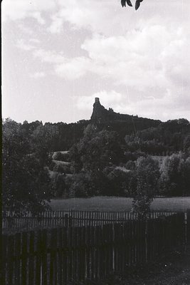 Rustic, weathered wooden fence in the foreground frames a view of a dramatic, ruined castle perched atop a heavily wooded hil...
