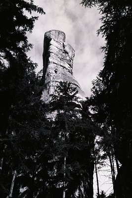 Towering rock formation rises dramatically amidst dark coniferous forest. Distinct layers and weathering patterns visible on ...